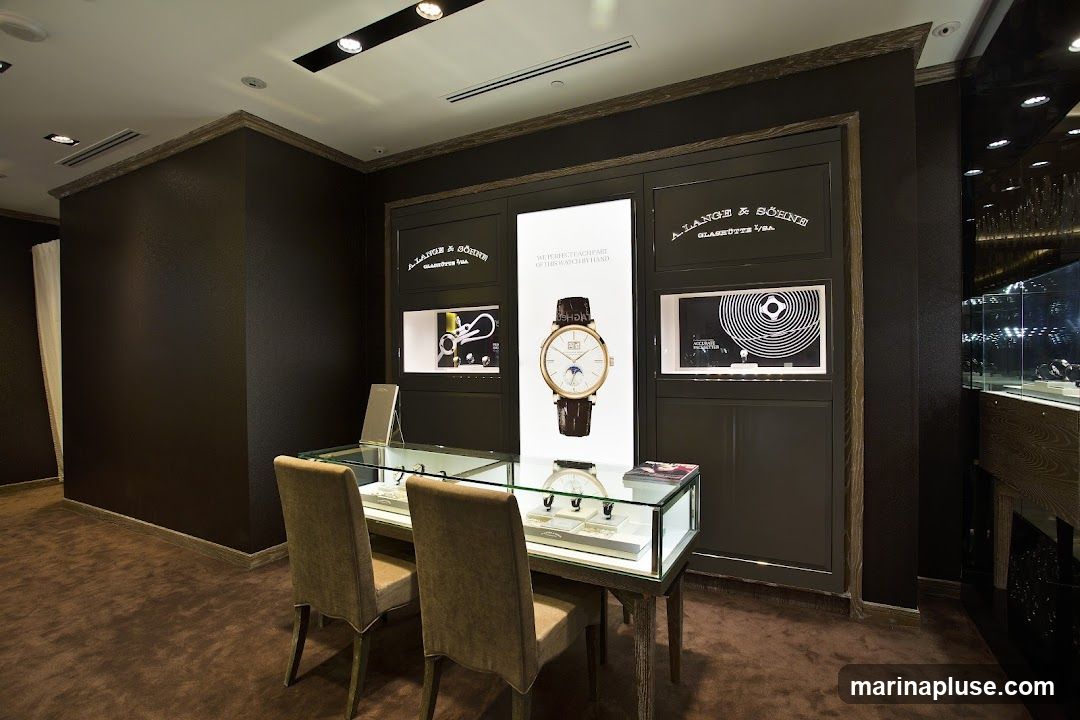 An interior view of a luxury watch store, featuring a display of A. Lange & Söhne timepieces. The central display showcases a large advertisement for a watch with the text "WE PERFECT EACH PART OF THIS WATCH BY HAND." Flanking this are smaller display cases with watches and brand logos. In the foreground, a glass-topped display table with two chairs in front of it holds more watches. The store has dark walls, brown carpeting, and elegant wooden accents.