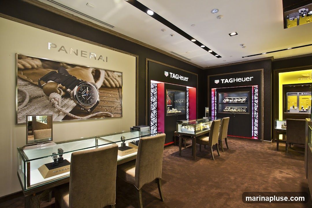 The interior of a high-end watch store featuring displays for Panerai, TAG Heuer, and Breitling watches. The store has plush carpeting, elegant display cases filled with watches, and large promotional images of watches on the walls. Seating is provided for customers.