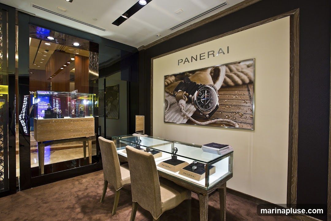 A luxurious interior display for Panerai watches. A large advertisement featuring a Panerai watch is mounted on the wall. In the foreground, a glass display case showcases several watches on stands, with two chairs positioned in front of it. Another display case is visible in the background.