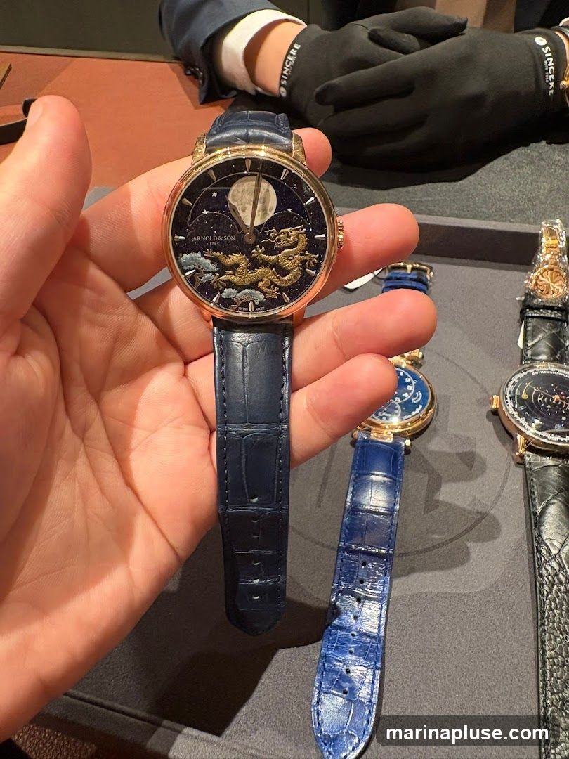 A person is holding a luxury watch with a dark blue alligator strap. The watch face is dark blue with a moon phase complication and features a golden dragon motif with celestial elements. The brand name "ARNOLD & SON" and "1764" are visible on the dial. In the background, other watches with similar straps are displayed, and a person wearing black gloves and a suit is visible.