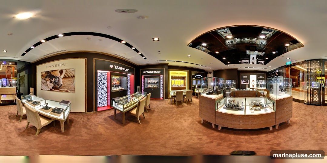A panoramic view of a high-end watch store. Display cases filled with watches are arranged throughout the store, with seating areas for customers. Prominent brand signage for Panerai and TAG Heuer is visible on the walls and displays. The store features plush carpeting and elegant lighting.