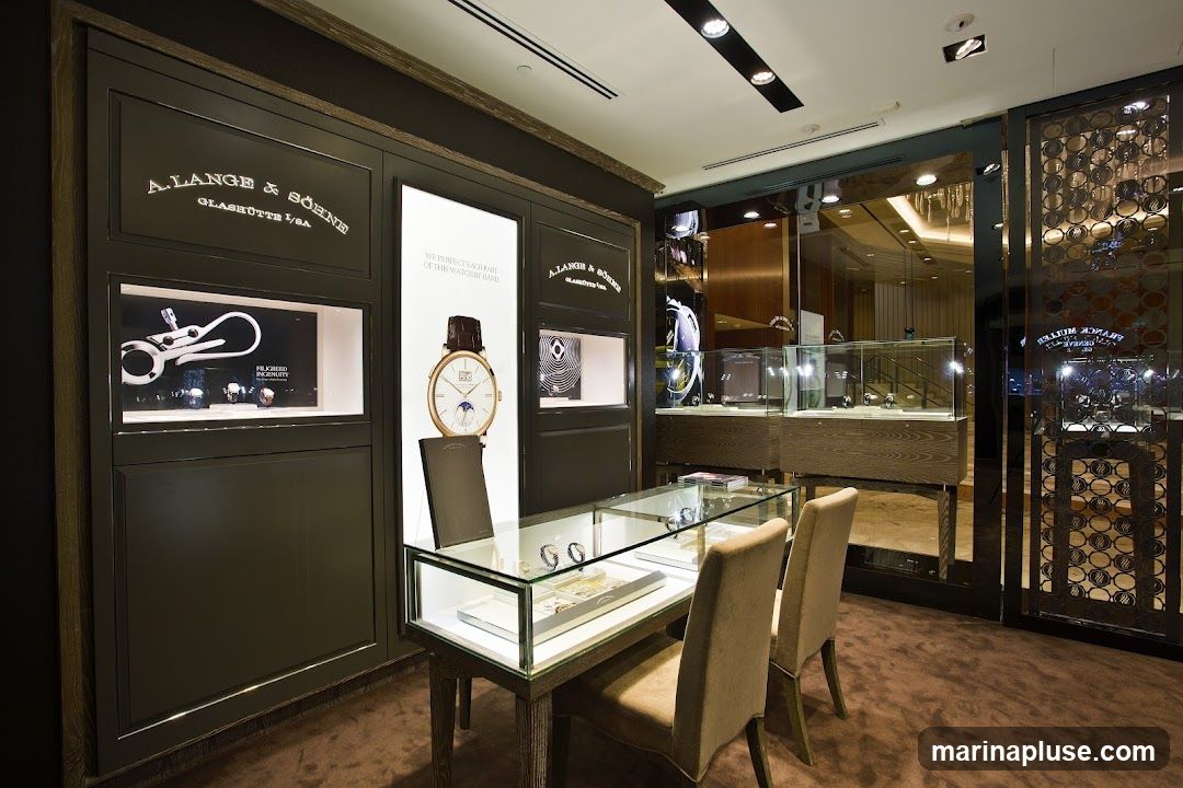A well-lit interior display of luxury watches. The main display features the A. Lange & Söhne logo and branding, with illuminated showcases displaying watches and watch components. A large poster showcases a detailed watch face with a moon phase complication. A glass display case in the foreground holds several watches, with two chairs positioned for customers. The background shows more display cases and a mirrored wall reflecting the store's interior.