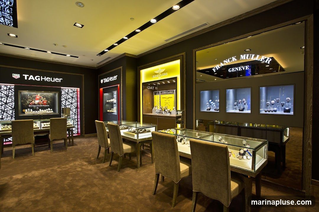 The interior of a high-end watch store featuring displays of TAG Heuer, Breitling, and Franck Muller timepieces. The store has plush carpeting, elegant display cases filled with watches, and comfortable seating for customers. The lighting is sophisticated, highlighting the luxury products.