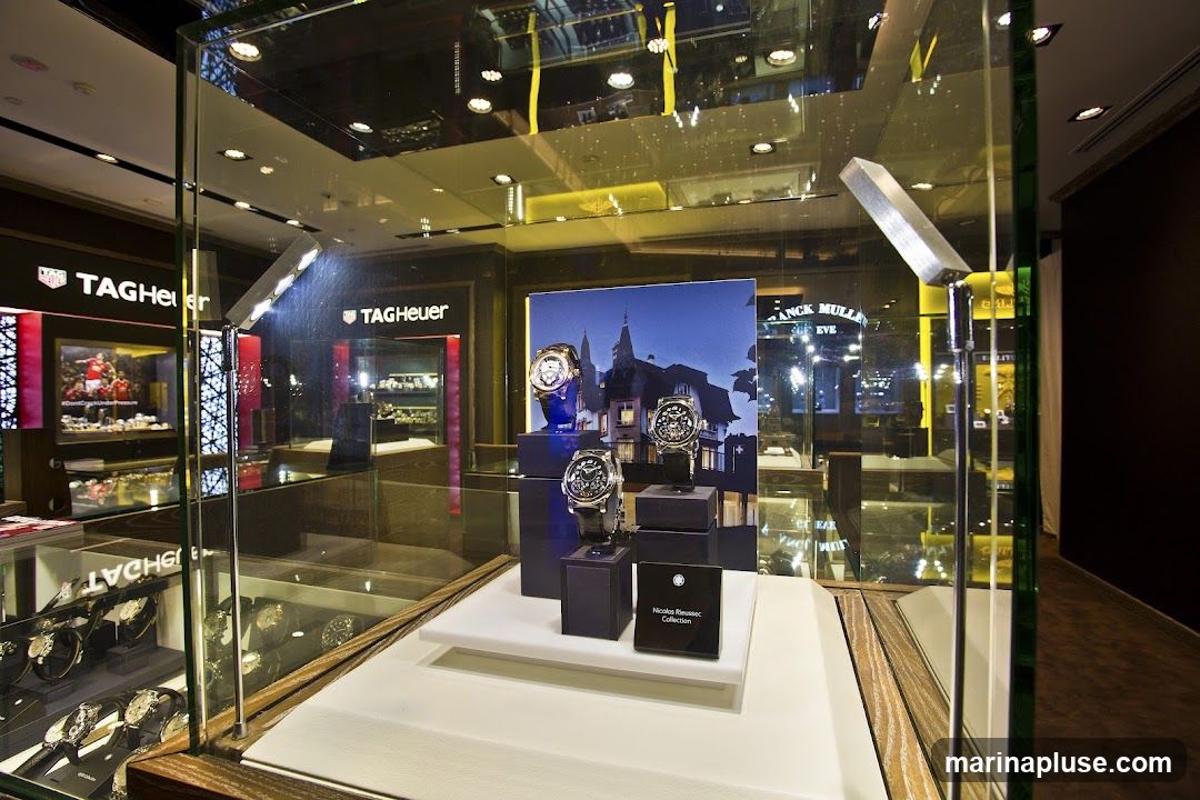 A display case showcasing luxury watches. In the foreground, three watches are presented on dark blue stands against a backdrop featuring a building and a night sky. The watches appear to be from the "Nicolas Rieussec Collection." In the background, the interior of a store is visible, with prominent "TAG Heuer" signage and displays of more watches.