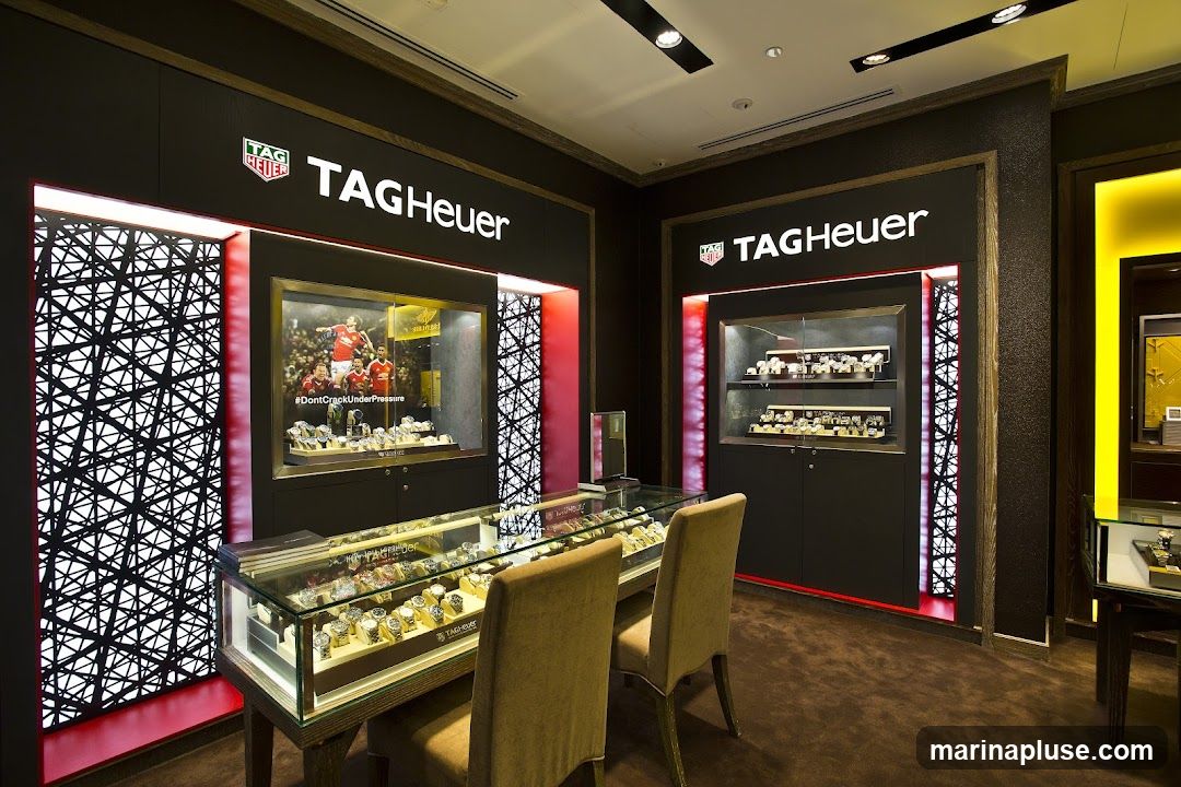 The interior of a TAG Heuer watch store, featuring display cases filled with watches. One display case shows a promotional image of soccer players with the hashtag #DontCrackUnderPressure. The store has modern lighting and dark, luxurious finishes.