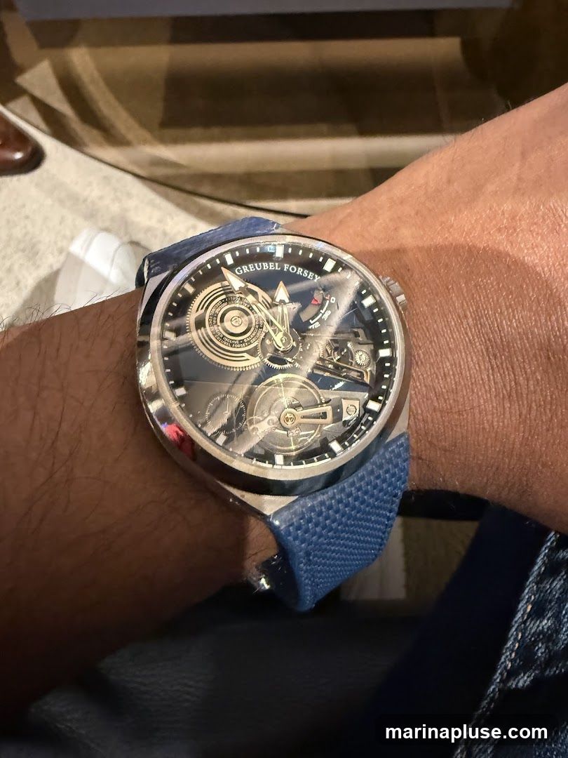 A close-up shot of a Greubel Forsey watch on a person's wrist. The watch has a complex mechanical dial with visible gears and components. The watchband is blue and textured. The background is blurred, showing hints of furniture and clothing.