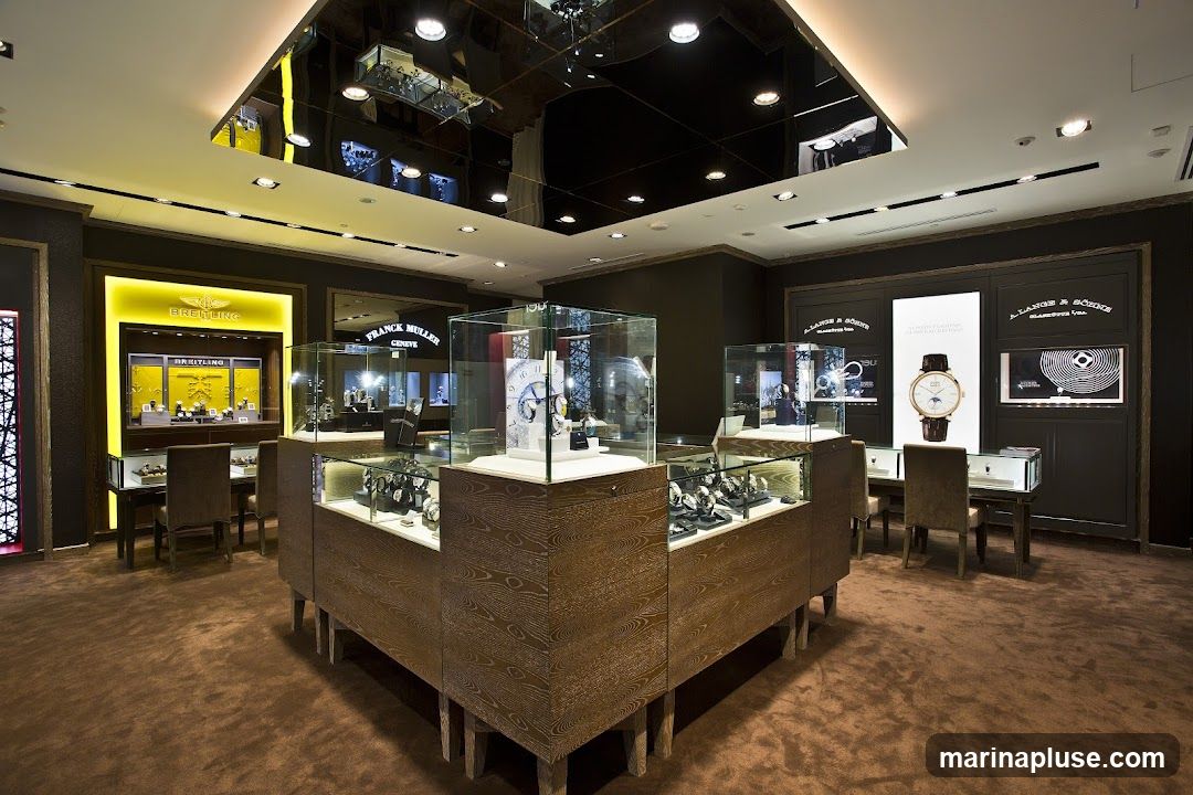 The interior of a high-end watch store, featuring display cases filled with watches. Prominent brands like Breitling and Franck Muller are visible on signage and displays. The store has a luxurious feel with dark wood accents, plush carpeting, and modern lighting.