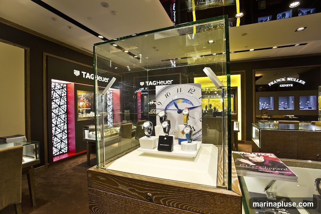 A well-lit interior of a luxury watch store, featuring prominent displays of TAG Heuer and Franck Muller timepieces. A central glass display case showcases several watches on white stands, with a large, artistic clock face in the background. Other display cases and shelves are visible, stocked with various watches. A magazine featuring a person on the cover is placed on a counter in the foreground.