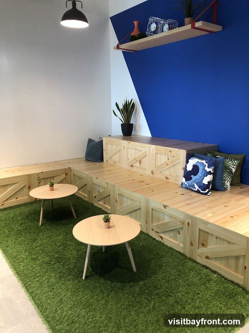 A contemporary seating area featuring tiered wooden benches with decorative paneling, two small round wooden tables, and artificial grass flooring. Several decorative pillows with wave and geometric patterns are placed on the benches. A snake plant in a black pot sits on the top bench, and a shelf above displays decorative items. The wall behind the seating area is painted with a striking blue and white geometric design. A black pendant light hangs from the ceiling.
