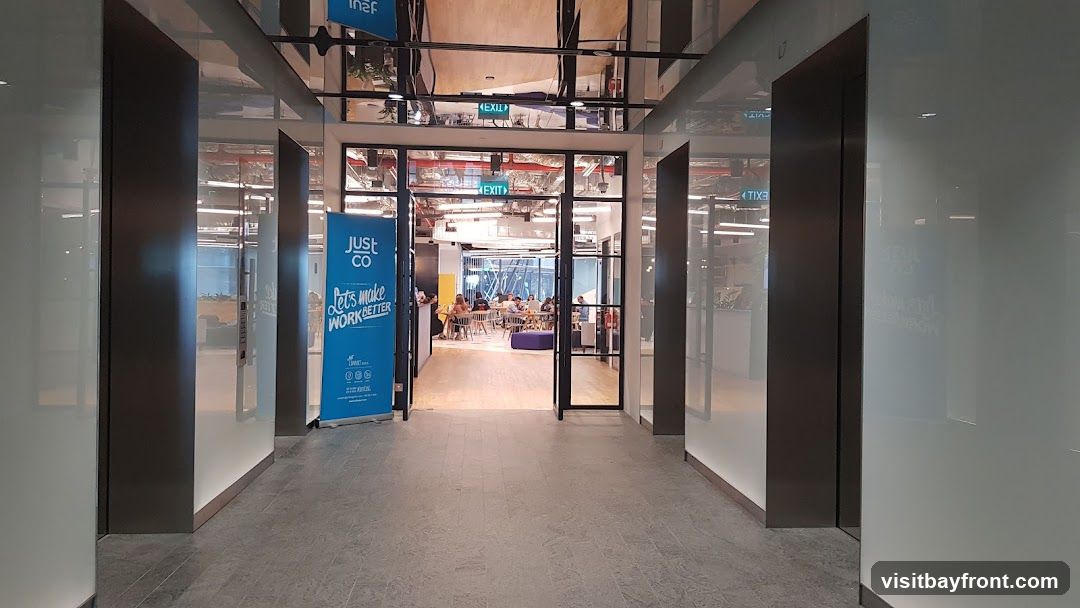 A view down a hallway towards an open office space. On the left, a blue banner displays "JUST CO Lets make WORK BETTER" with social media icons. The office interior features tables and chairs, with people seated and working. The ceiling has a reflective surface, showing the office lights and infrastructure. "EXIT" signs are visible.