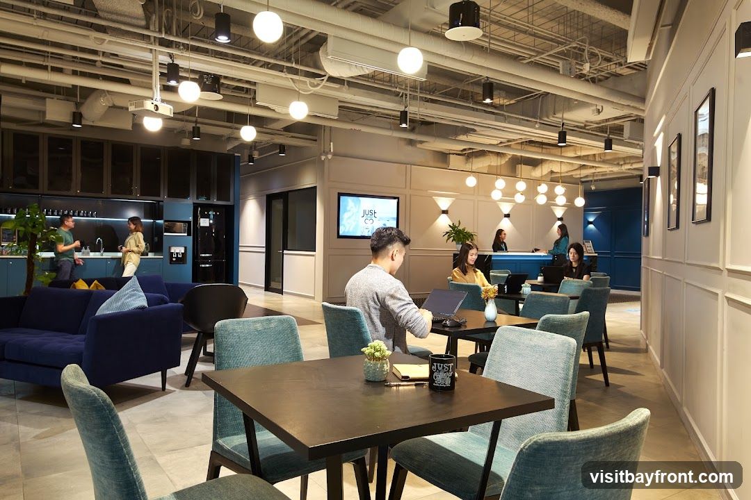 A spacious and modern co-working space with comfortable seating areas, including sofas and tables with chairs. People are seen working on laptops and interacting. A kitchen area with a refrigerator and water dispenser is visible in the background. The space is well-lit with various types of lighting fixtures. A screen displays the logo "just go".