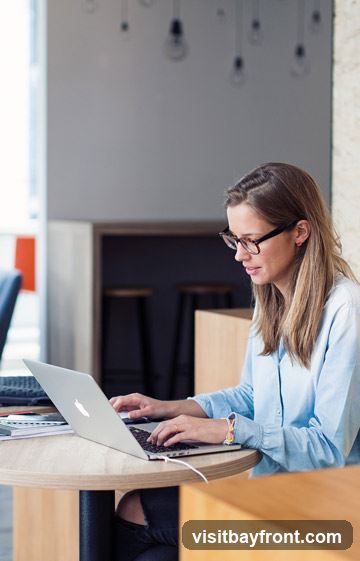 A woman with glasses and long blonde hair is sitting at a round wooden table, typing on a silver laptop. She is wearing a light blue collared shirt. The background shows a modern interior with hanging light bulbs and wooden accents.