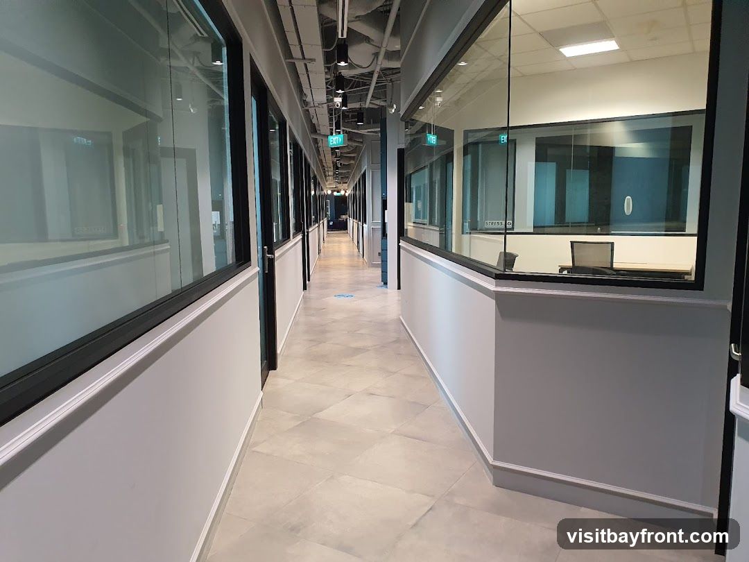 A modern office corridor with glass-walled rooms on either side. The floor is tiled with a diamond pattern. The ceiling has exposed pipes and lighting fixtures. An exit sign is visible in the distance. One room contains a desk and chair.