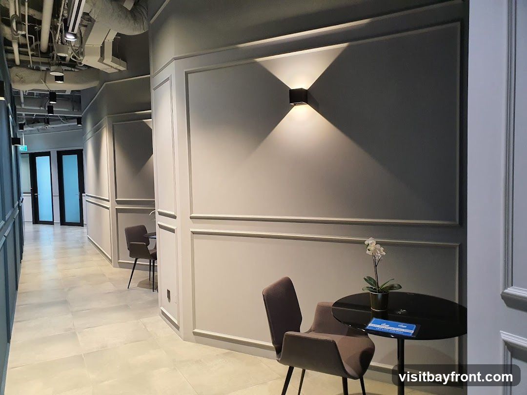 A well-lit hallway in a modern office space. The walls are painted a neutral gray with decorative molding. A contemporary wall sconce casts a warm glow. In the foreground, a small round table with a modern chair holds a potted orchid and a blue sign. The floor is tiled with large, light-colored tiles. In the background, a corridor leads to double glass doors with an "EXIT" sign. Exposed pipes and ductwork are visible on the ceiling.
