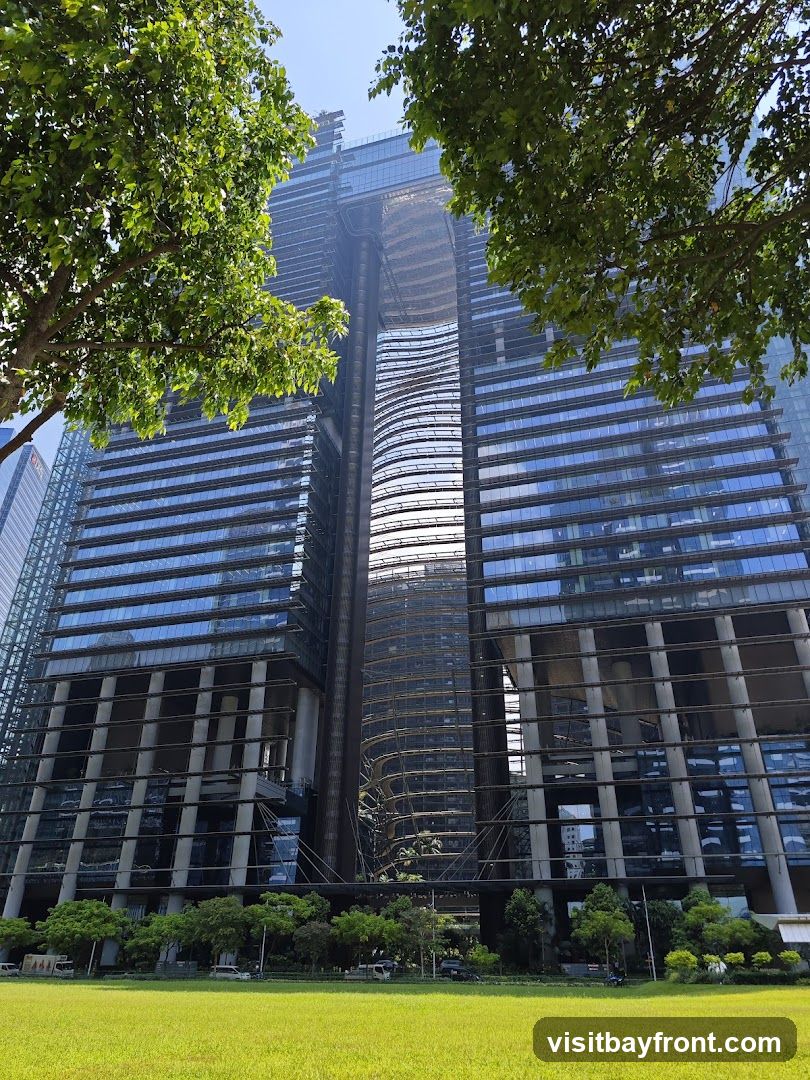A low-angle shot of a large, modern skyscraper with a unique architectural design, featuring a central open section and curved glass facades. The building appears to be under construction, with scaffolding visible in the central void. Lush green trees frame the top of the image, and a manicured lawn with a few cars is in the foreground. The sky is clear and blue.