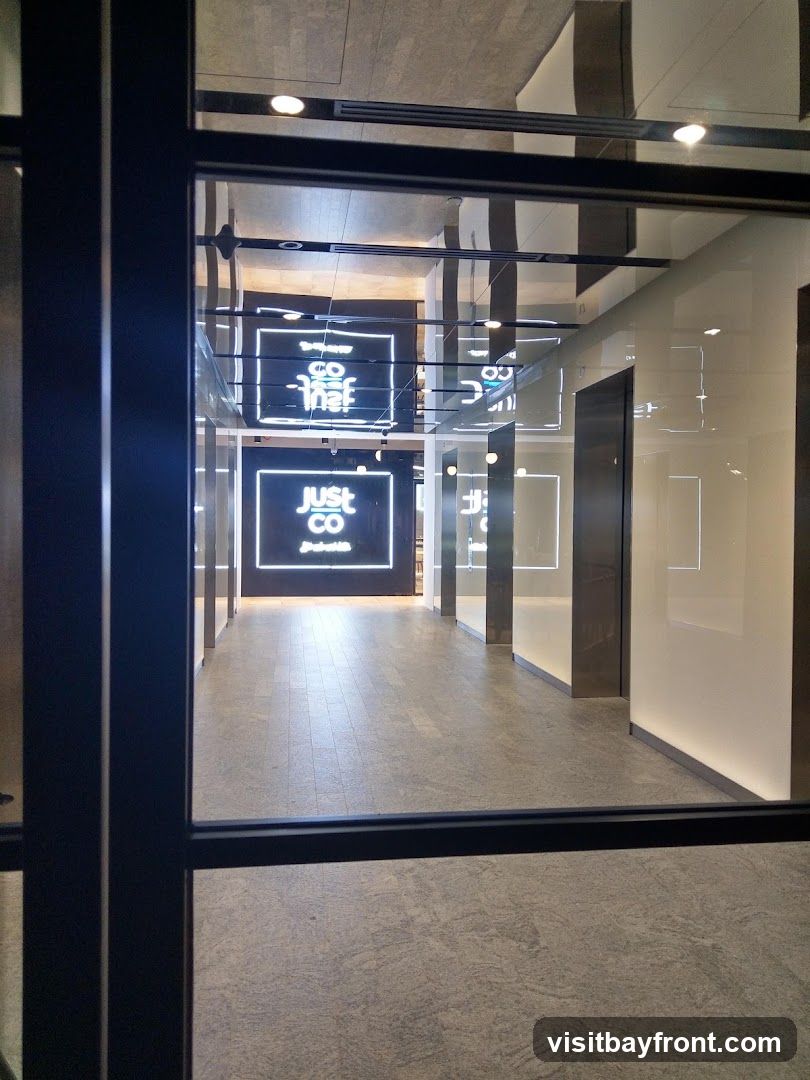 A view through a glass partition into a modern interior hallway. The hallway has a tiled floor and illuminated walls. Several square neon signs with the text "just CO" are visible, some with additional text below. The signs are reflected in the polished ceiling.