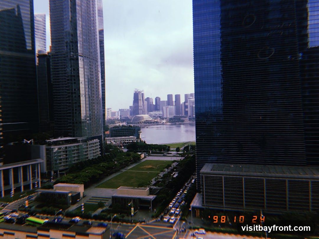 A panoramic view of a modern cityscape, dominated by tall skyscrapers. In the foreground, a large green field is surrounded by trees and pathways. A body of water, possibly a bay or river, separates the foreground from a distant skyline of more buildings. Cars are visible on a road in the lower part of the image. A date stamp '98 10 24' is visible in the bottom right corner.