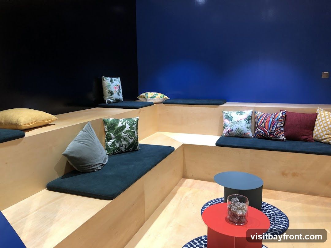 A contemporary seating area with tiered wooden benches covered in dark teal cushions. The benches are adorned with an assortment of decorative pillows featuring tropical leaf prints, abstract patterns, and solid colors like burgundy and yellow. The walls are painted in contrasting dark black and vibrant royal blue. In the foreground, two modern, colorful coffee tables, one red and one dark gray, are placed on patterned rugs. A glass vase filled with pinecones sits on the red table.
