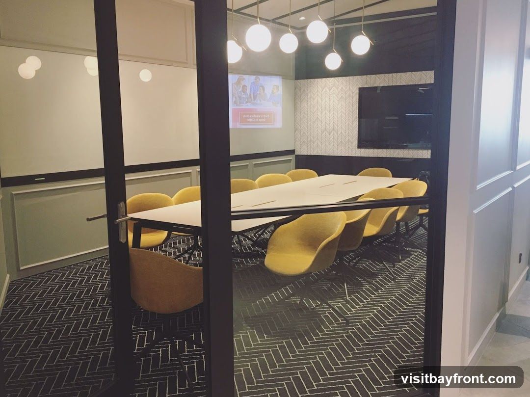 A view into a modern conference room with a long white table surrounded by yellow chairs. A projector screen displays an image of people, and a large flat-screen TV is mounted on the wall. The room has a patterned carpet and contemporary lighting fixtures.
