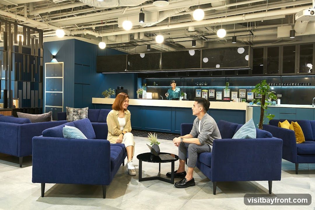 Two people are sitting on navy blue sofas in a modern co-working space. The woman on the left is wearing a beige cardigan over a white top and white pants. The man on the right is wearing a grey cardigan over a white t-shirt and grey pants. They are facing each other and smiling. A black coffee table with a vase of dried flowers sits between them. In the background, a woman in a green shirt is standing behind a counter, and there are shelves with various items and plants. The space has a modern design with exposed ceilings, pendant lights, and blue accent walls.