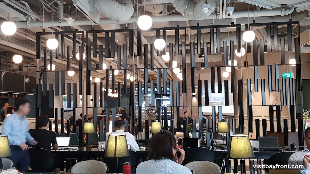 A spacious and modern co-working space with numerous individuals working on laptops. The interior features a striking decorative partition made of vertical black slats and mirrored panels, interspersed with numerous globe pendant lights. Desks are arranged in rows, each equipped with lamps and laptops. An 'EXIT' sign is visible in the background.