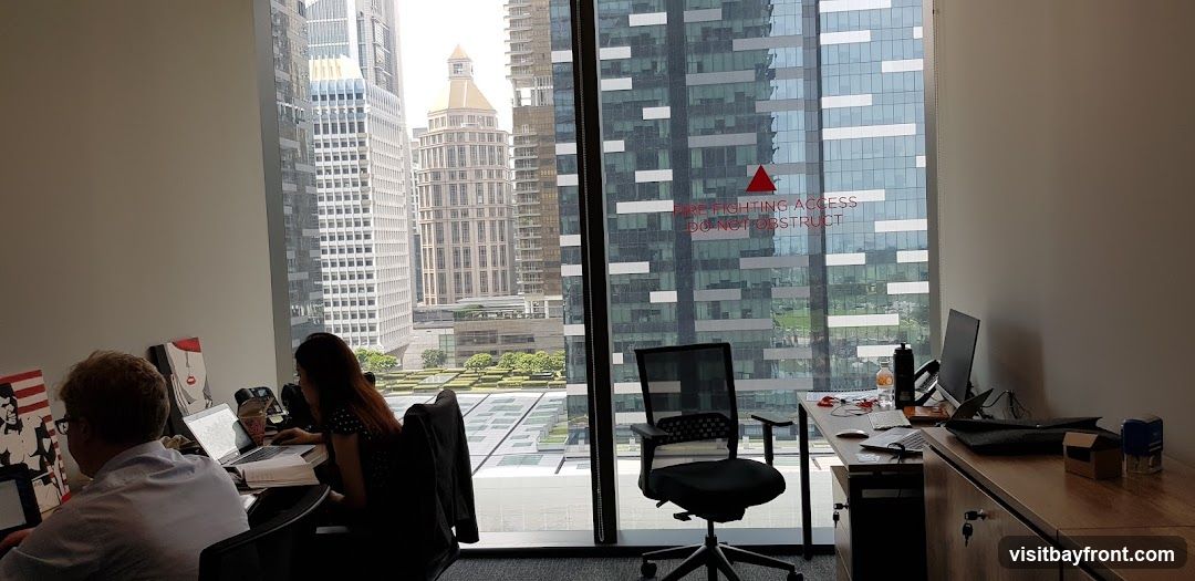 An office interior with two people working at desks. The desks are positioned in front of a large window that offers a panoramic view of a city skyline with tall buildings. One person is a man with glasses, facing away from the camera, working on a laptop. The other person is a woman, also working on a laptop. Artwork is displayed on the wall behind the man. The window has a red triangular sticker with text that reads "FIRE FIGHTING ACCESS DO NOT OBSTRUCT".