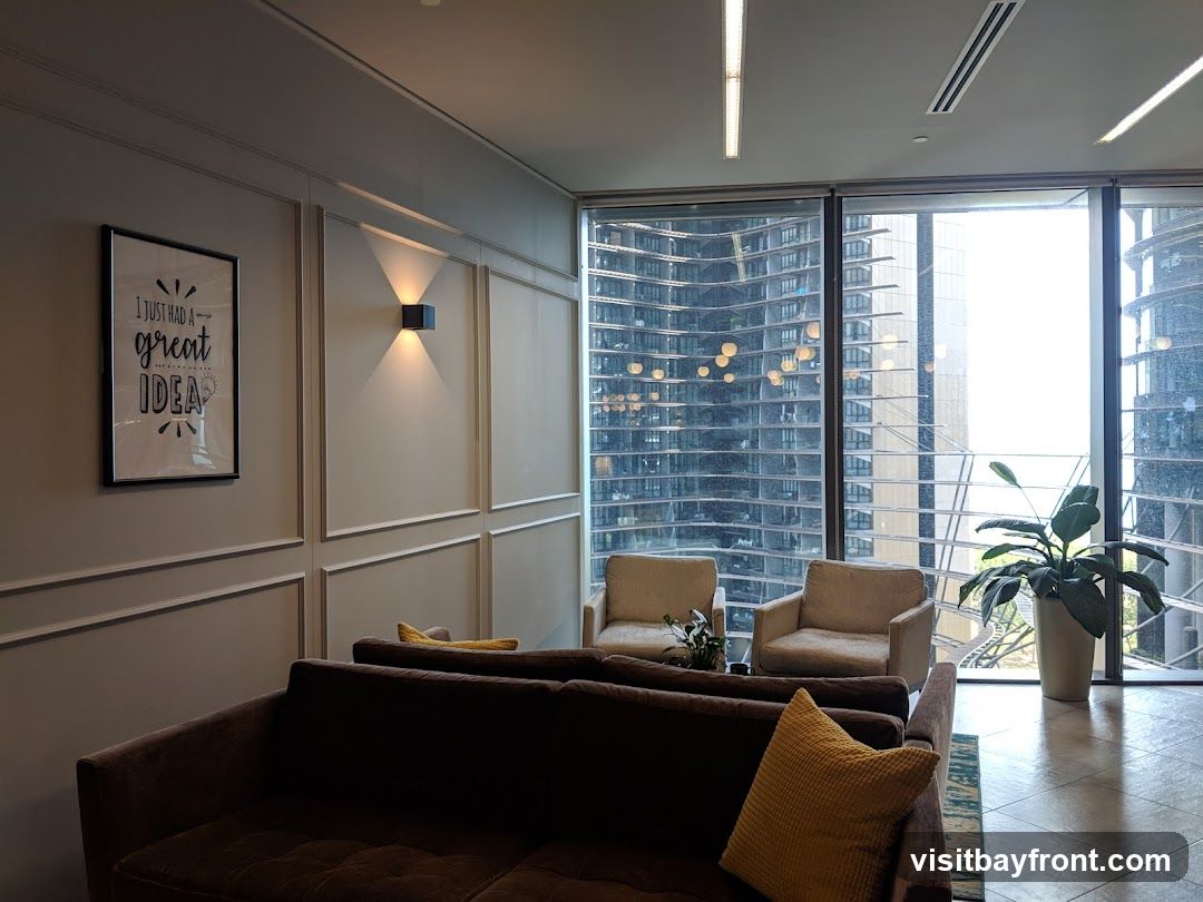 A contemporary lounge area featuring a dark brown tufted sofa with yellow accent pillows, two beige armchairs, and a large potted plant. The room has a framed print on the wall that reads "I JUST HAD A great IDEA" and a modern wall sconce. Large windows offer a view of a dense urban cityscape with modern architecture.