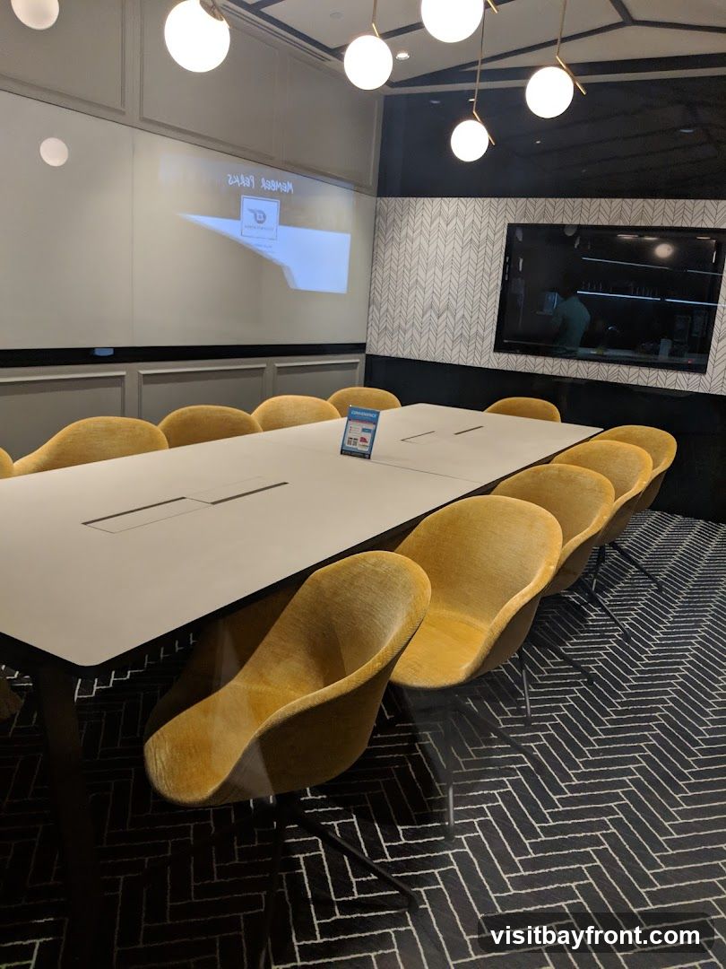 A modern conference room with a long table and yellow chairs. A projector screen displays a logo and text, and a large monitor is mounted on the wall.
