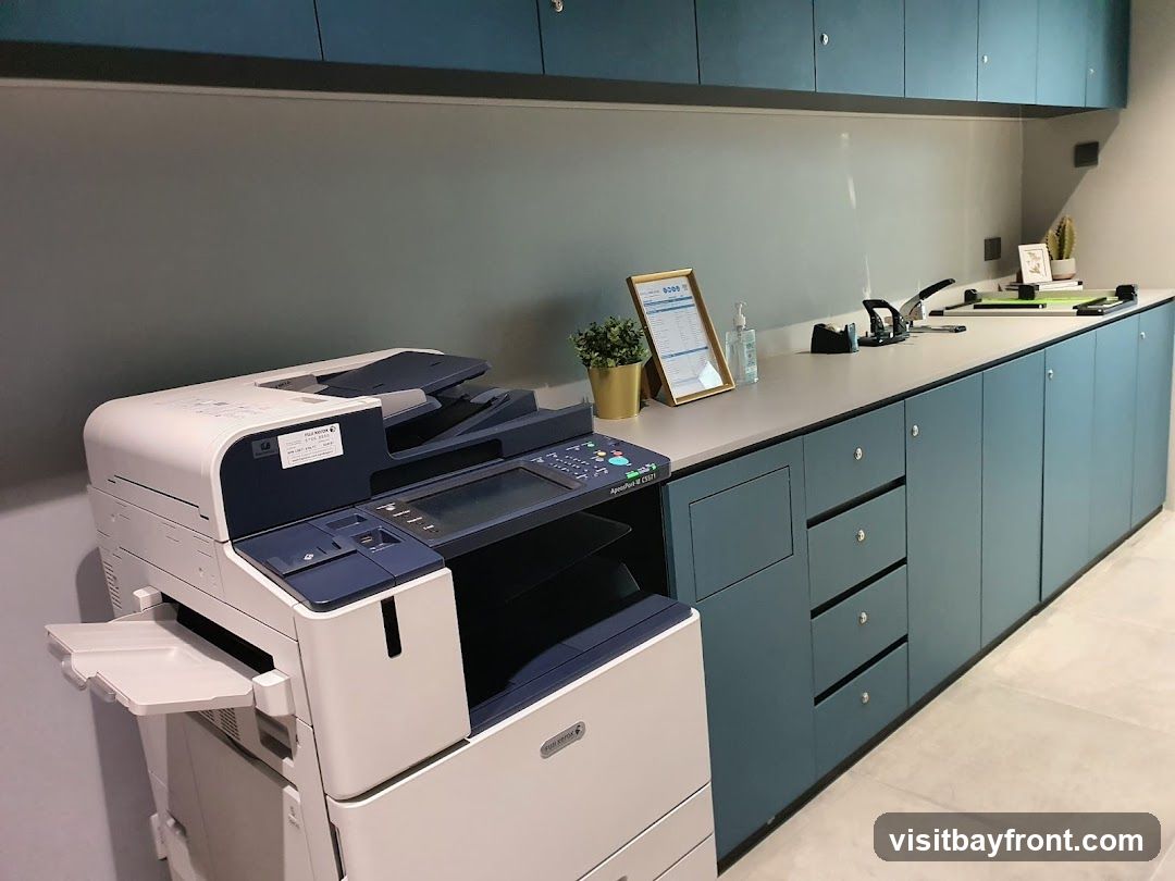 A modern office space featuring a large Fuji Xerox copy machine on the left. To the right, a long countertop displays a framed document, a bottle of hand sanitizer, two hole punches, and other office supplies. The counter is lined with teal cabinets and drawers.