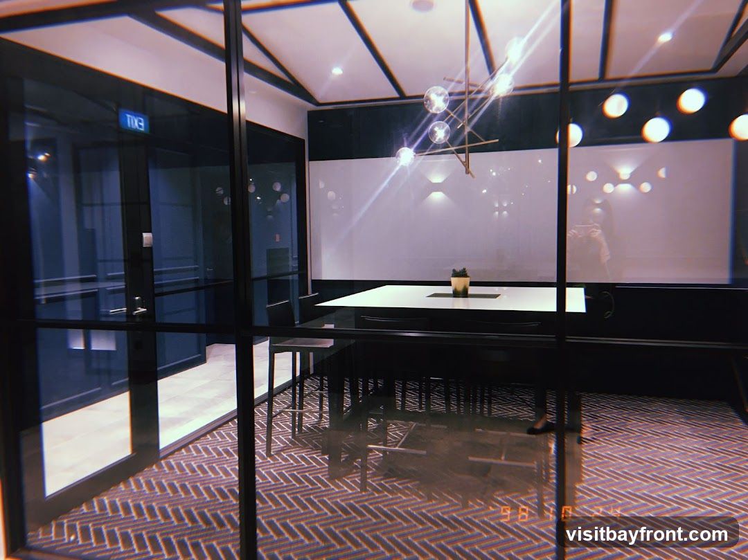 A modern meeting room with glass walls, a large table, and chairs. The room has a geometric chandelier and recessed lighting. The floor is carpeted with a herringbone pattern. An exit sign is visible on the glass door.