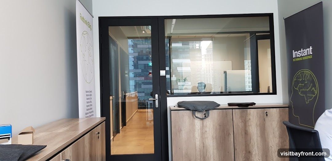 An office interior featuring a glass-paneled door leading to a hallway. Two large banners are visible: one on the left with a globe graphic and text, and another on the right with the text "Instant Rethinking Workspace" and a brain graphic. Wooden cabinets are present along the walls. Through the glass, a person is visible working at a desk in a well-lit room with whiteboards.