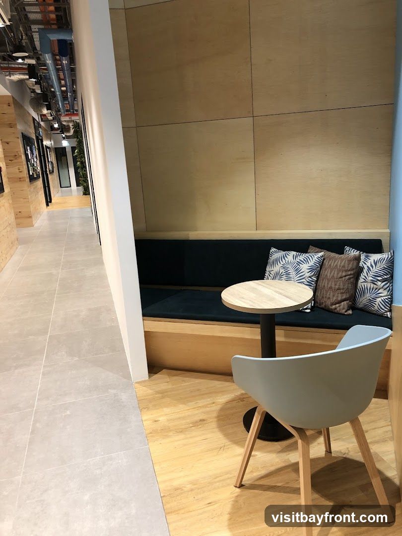 A contemporary seating area with a built-in dark blue bench, wooden wall panels, and a light blue chair. A small round table with a wooden top sits in front of the bench. The area is adjacent to a tiled hallway with exposed ceiling pipes and lighting fixtures.