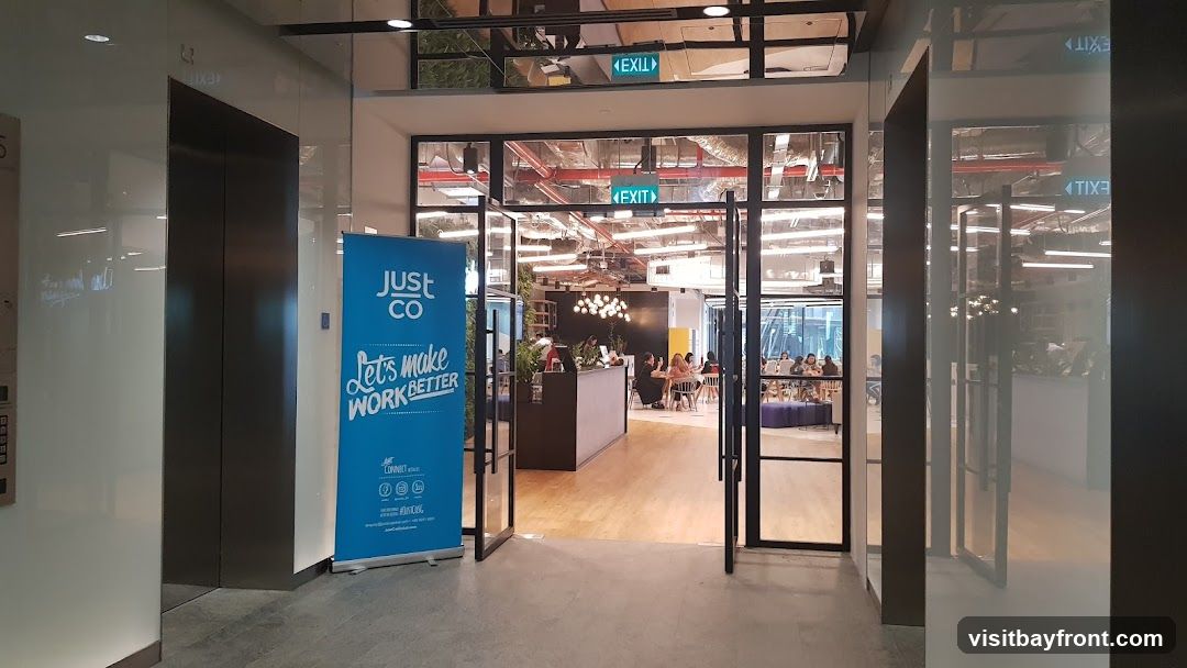 An open doorway leads into a bright, modern co-working space. A blue banner with the text "JUST CO Let's make work BETTER" stands to the left of the entrance. Inside, people are seated at desks and tables, working on laptops. The space features exposed ceilings with industrial lighting and a contemporary chandelier.