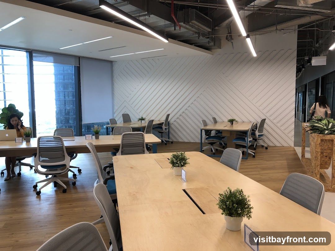 An interior view of a modern co-working space with multiple desks and chairs. A woman is seated at a desk on the left, using a laptop and talking on her phone. The space features large windows, wooden floors, and a decorative white wall with a geometric pattern.