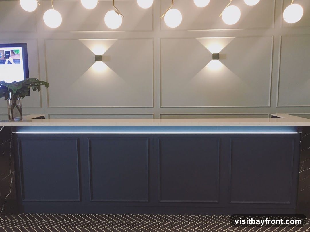 A sleek, dark blue reception desk with a white countertop is illuminated by subtle LED lighting. The wall behind the desk features elegant paneling and two modern wall sconces casting warm light. A glass vase with a large green plant sits on the left side of the counter. A digital screen is visible on the far left, displaying what appears to be a welcome message. The floor has a black and white herringbone pattern.