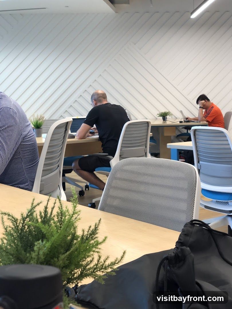 Several people are seated at desks in a contemporary office space, working on laptops. The walls have a distinctive geometric pattern. One person in the foreground is wearing a checkered shirt, while another in the mid-ground wears a black t-shirt and shorts. Further back, a person in an orange shirt is also working at a laptop. The desks are made of light wood, and modern office chairs are visible. A small potted plant sits on one of the desks.