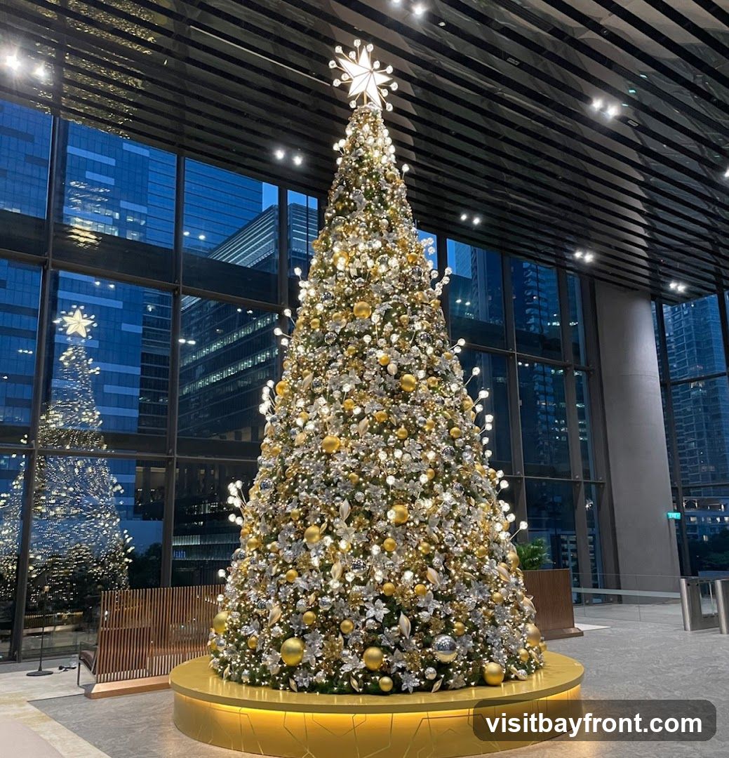 A large, elaborately decorated Christmas tree stands in a modern lobby with large windows reflecting city buildings. The tree is adorned with gold and silver ornaments, lights, and star-shaped accents. A smaller, reflected Christmas tree is visible in the window.