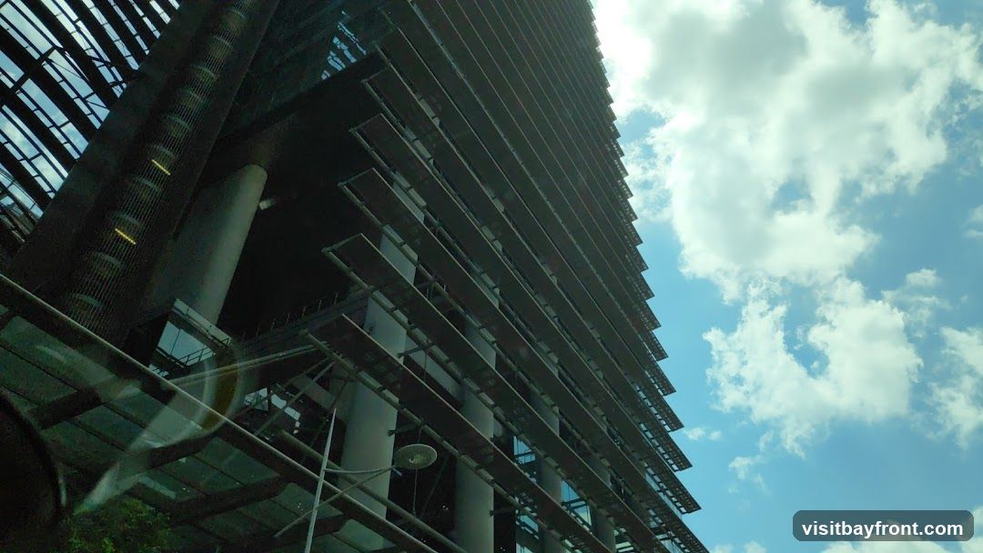 A low-angle shot of a modern skyscraper with a distinctive facade featuring horizontal louvers and glass panels. The building's structure is visible, with large cylindrical columns supporting upper levels. The sky above is bright blue with scattered white clouds.