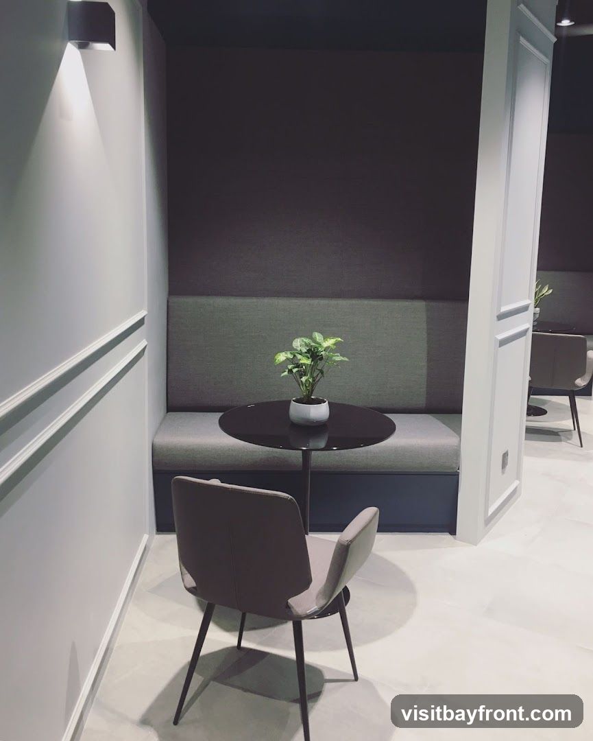 A modern seating area with a dark round table, a potted plant, and upholstered chairs and banquette seating. The walls are light gray with decorative molding, and a wall-mounted light fixture illuminates the space. The floor is tiled with a light marble pattern.