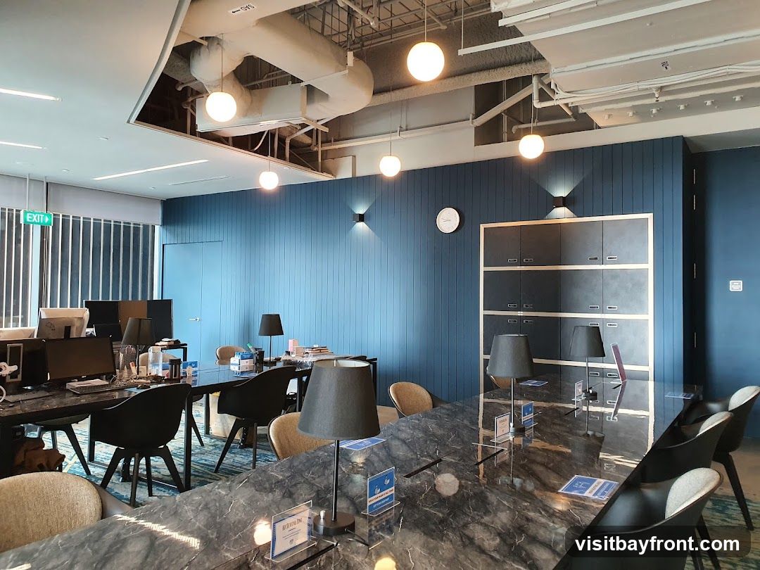 An interior view of a modern office space with blue paneled walls, large marble-topped tables, and black chairs. Desks with computers are visible on the left, and a wall of cabinets is on the right. Pendant lights and wall sconces illuminate the room. An exit sign is visible on the left.