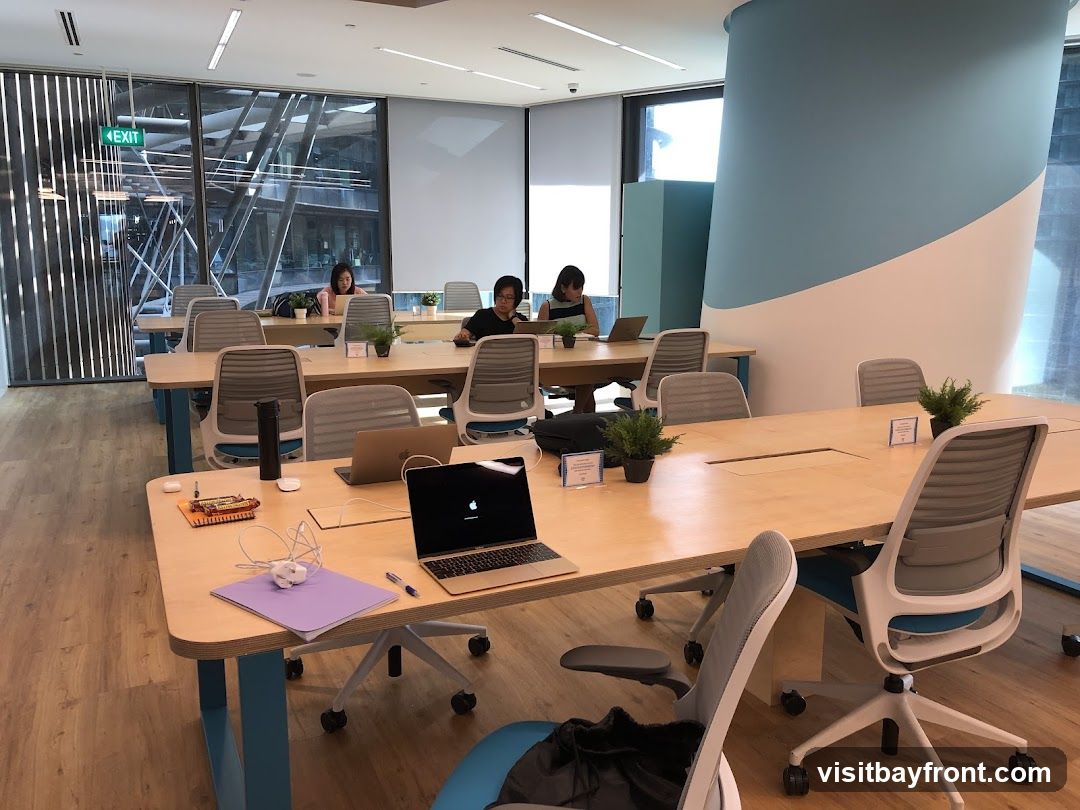 An interior view of a contemporary co-working space with multiple desks and chairs. Several individuals are seated at the desks, working on laptops. Large windows offer a view of an urban landscape. The space features light wood furniture, blue accents on the furniture and a large curved pillar, and potted plants on the tables. An "EXIT" sign is visible near the window.