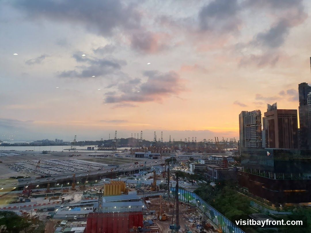 An aerial view of a busy port and construction site at sunset. The sky is filled with soft orange, pink, and purple hues. In the foreground, there is a construction site with cranes and buildings under development. Beyond that, a large port area is visible with many shipping containers and cranes. In the distance, a city skyline can be seen.