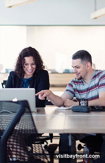A woman with curly dark hair and a man with short dark hair are sitting at a table, looking at a laptop. The woman is wearing a dark blazer over a blue shirt, and the man is wearing a striped t-shirt. The man is pointing at the laptop screen. There is a black mug with the word "JUST" on it, a pair of glasses, and a phone on the table. The background appears to be an office setting with modern lighting fixtures.