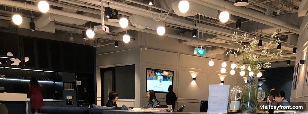 An interior view of a modern office break area with various seating arrangements and decorative elements. The ceiling features exposed pipes and a projector, with numerous pendant lights and wall sconces providing illumination. A large screen displays colorful images, possibly a menu or promotional content. Several individuals are seen interacting or working in the space. An illuminated green 'EXIT' sign is visible on the ceiling.