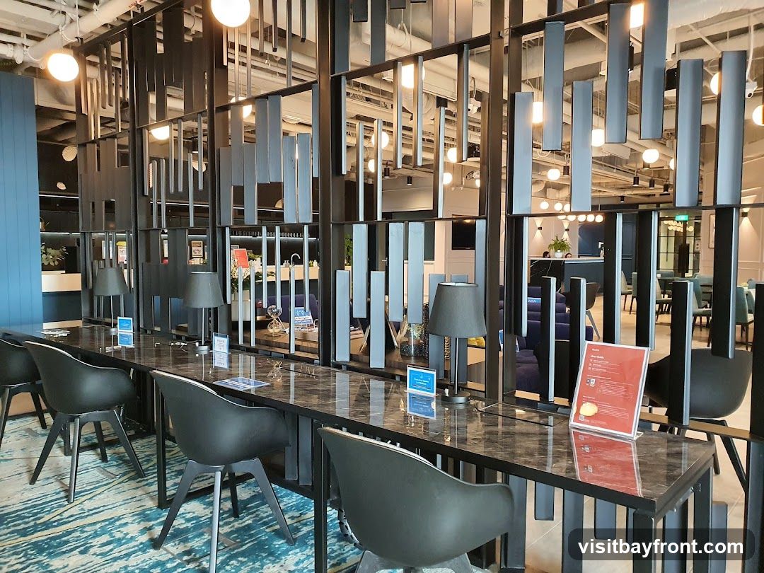 A long, polished desk with black chairs in a contemporary co-working space. The desk is equipped with lamps and small informational signs. Behind the desk, a decorative screen separates the area from a lounge with plush seating and a reception desk in the background. The space features modern lighting and a patterned carpet.
