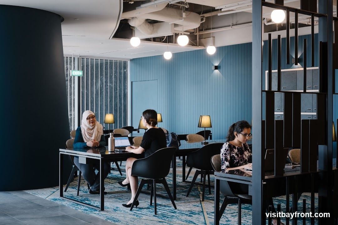 A contemporary co-working space with a blue paneled wall and modern lighting. Three women are seated at tables, engaged with laptops. One woman wears a hijab and glasses, another has her back to the camera, and the third is focused on her laptop. The space features sleek black tables and chairs, with decorative lamps on some tables. A blue and white patterned rug covers the floor. An exit sign is visible on the left side of the image.