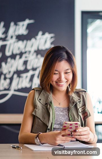 A young woman with short brown hair smiles as she looks down at her smartphone. She is wearing a grey patterned top and a green utility vest with patches. She is sitting at a wooden desk with a notebook, pen, and watch. Behind her, a dark wall has white chalk writing on it.