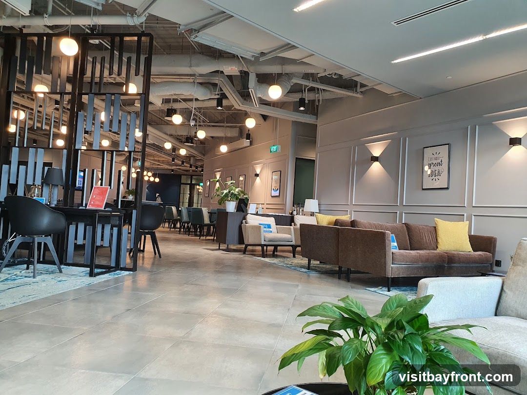 A spacious and modern co-working space with comfortable seating areas, desks, and decorative lighting. The interior features a mix of contemporary furniture, including sofas, armchairs, and office chairs, arranged around tables. The walls are adorned with framed artwork and wall sconces, creating a warm and inviting atmosphere. A large plant in the foreground adds a touch of greenery to the scene.