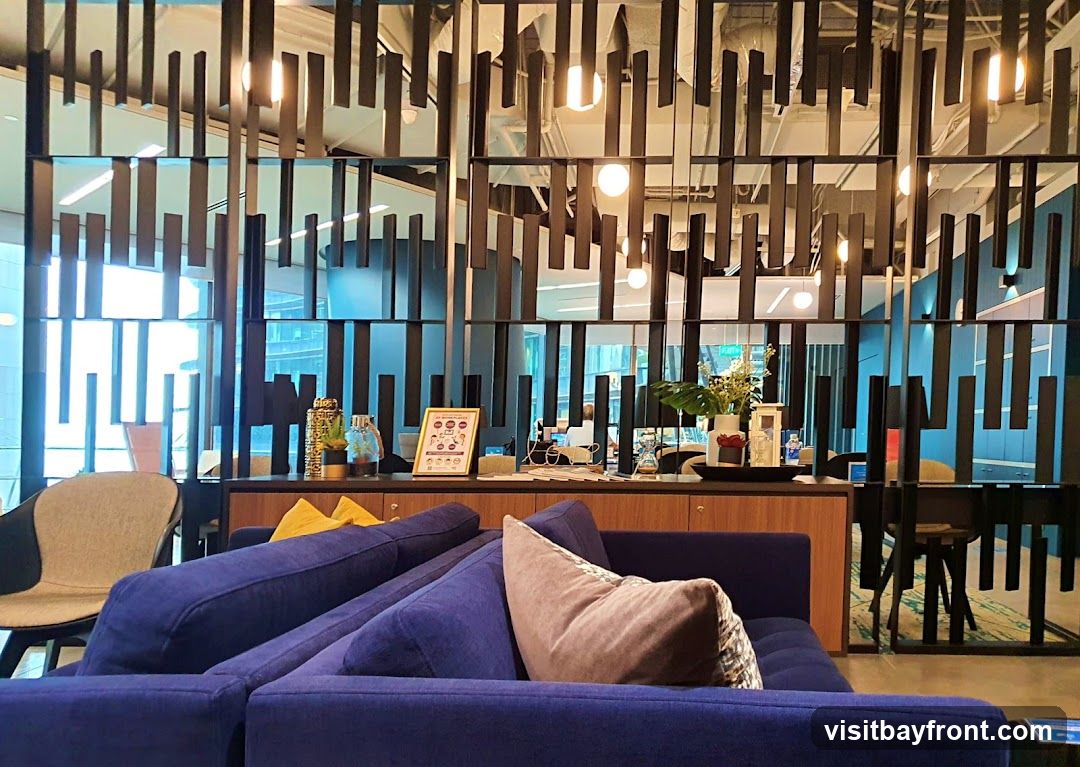 A contemporary lounge area with a large blue sectional sofa in the foreground. Behind the sofa is a wooden counter with various decorative items, including vases, plants, and a framed poster. A modern partition with vertical black slats separates the lounge from a workspace visible in the background. Natural light streams in from large windows.