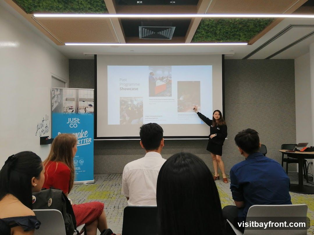 A woman is presenting a slideshow titled "Past Programme Showcase" to an audience. The presentation includes images and text descriptions of past programs. There are banners with the logo "JUST CO" and the slogan "make BETTER" visible on the left side of the room. The room has a modern design with green ceiling panels and white walls.